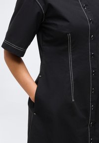 Μαύρο φόρεμα με κοντά μανίκια και λευκή αντίθεση ραφής, με κουμπιά μπροστά και πλαϊνές τσέπες, ένα χέρι ακουμπισμένο μέσα στην τσέπη.