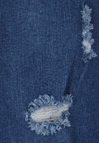 Kék farmeranyag, amely vonzó kifakult foltokkal és elhasználódott részekkel rendelkezik. Különösen a szakadt részek körül látható kopás, amely textúrált felületet mutat.