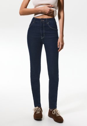 Frau trägt dunkelblaue High-Waist-Skinny-Jeans, beige ärmelloses Oberteil und braune Schnürschuhe, steht vor einem weißen Hintergrund.