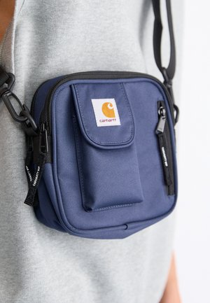 Marineblaue Carhartt Crossbody-Tasche mit Fronttasche und Reißverschluss, getragen über einem grauen ärmellosen Top.