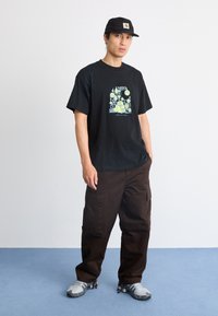 Sort bomulds T-shirt med et farverigt grafisk design. Kombineret med mørkebrune cargobukser og grå fodtøj. En simpel kasket fuldender looket.