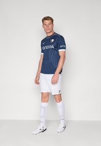 Camiseta de fútbol de la marina con acentos en blanco, con rayas verticales y un logo. Combinada con pantalones cortos blancos y calcetas blancas hasta la rodilla.