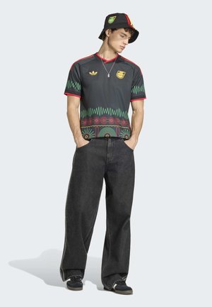 Jeune homme portant un t-shirt graphique noir, un jean large noir, des baskets noires, un chapeau avec des rayures rouges et jaunes, debout les mains dans les poches.