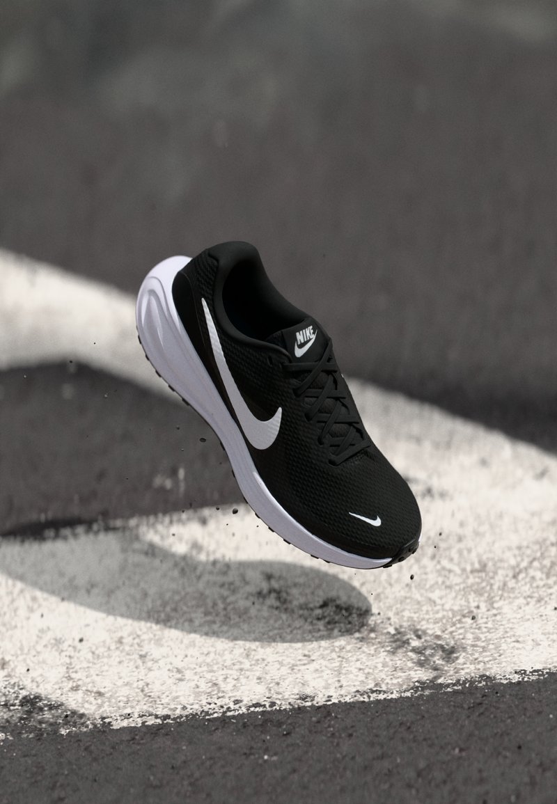 Chaussure de running Nike noire et blanche en l'air au-dessus d’un bitume avec marquages routiers blancs, soulevant petits graviers et poussière.