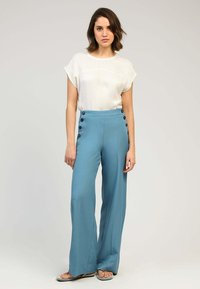 Pantalons bleus taille haute avec une jambe large et des accents de boutons noirs sur le côté, associés à un chemisier blanc à manches courtes.