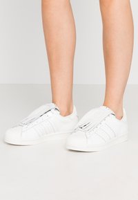 Ben med vita Adidas-sneakers med tre ränder och fransöverdrag över snörena på en enkel ljus bakgrund.