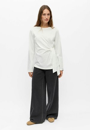 Femme debout portant un haut blanc à manches longues avec un nœud sur le côté, un pantalon large gris foncé et des chaussures plates beiges sur fond blanc.