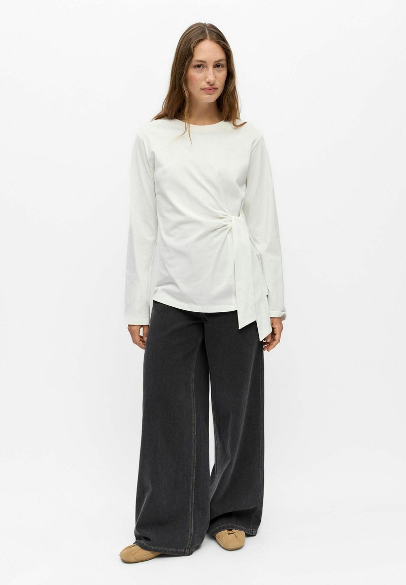 Femme debout portant un haut blanc à manches longues avec un nœud sur le côté, un pantalon large gris foncé et des chaussures plates beiges sur fond blanc.