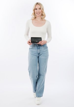 Mujer rubia que lleva una camiseta blanca de manga larga y jeans azules, sonriendo y sosteniendo una pequeña cartera negra frente a un fondo blanco.