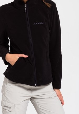 Person iført en sort fleecejakke med lynlås med den ene hånd i lommen og lyse cargobukser med forlomme.