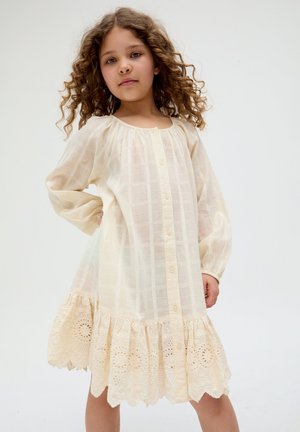 Junges Mädchen mit lockigem Haar, das ein cremefarbenes Kleid mit langen Ärmeln, Knopfleiste vorne und Spitzenabschluss trägt und vor einem schlichten hellen Hintergrund steht.