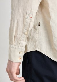 Camicia di lino beige con maniche lunghe, caratterizzata da bottoni in perla e un accento rosso. Orlo rotondo e etichetta laterale visibile vicino al fondo.