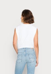 Mulher com cabelo castanho curto, vestindo uma blusa branca sem mangas e calças jeans de cintura alta azul claro, de costas contra um fundo simples.