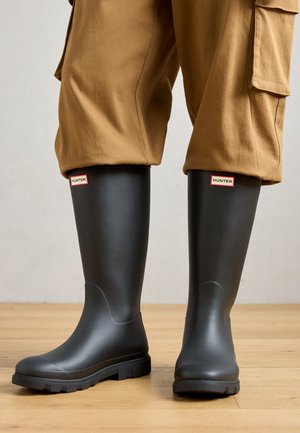 Persona wearing black Hunter rain boots and loose brown pants standing on a wooden floor against a plain background.
