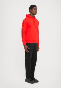 Sudadera roja con bolsillo canguro y logo negro en el pecho y la manga. Combinada con pantalones negros y zapatillas, sobre un fondo liso.