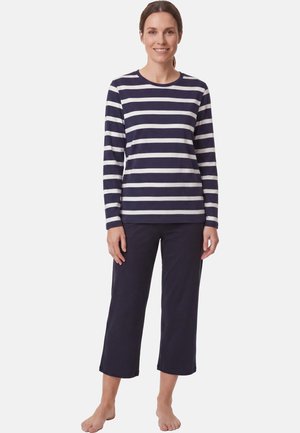 Frau steht barfuß und trägt ein marineblau-weiß gestreiftes Langarmshirt sowie marineblaue, verkürzte Hosen vor einem schlichten hellen Hintergrund.