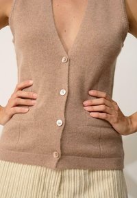 Jasnobrązowy bezrękawnik z głębokim dekoltem w kształcie V, z pięcioma guzikami, bocznymi kieszeniami i miękkim, teksturowanym wykończeniem.