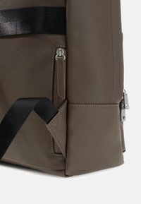 Mochila de tela texturizada marrón con un bolsillo con cremallera, correa negra y detalles en metal plateado. Diseño angular con costuras precisas.