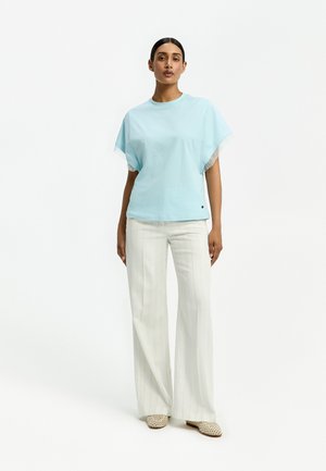 Frau steht, trägt ein hellblaues Kurzarmtop, weiße weite Hosen mit dezenten Streifen und beige gewebte Slipper vor weißem Hintergrund.