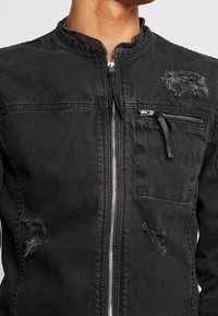 Musta farkkutakki hopeisella vetoketjulla, etupuolella vetoketjullinen rintatasku ja kuluneita kangaspäällysteitä rintakohdassa ja alaosassa.