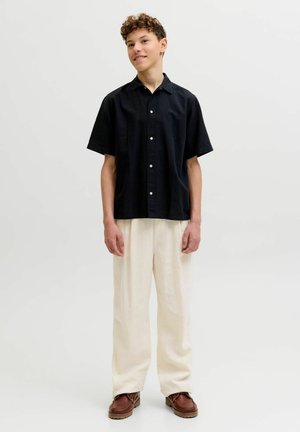 Fiatal személy áll, rövid ujjú fekete inget, bő fehér nadrágot és barna cipőt visel egy egyszerű világos háttér előtt.