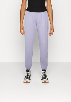 Person iført lyse lilla joggebukser med elastiske manchetter, sort top og sort-hvide høje sneakers mod en ensfarvet hvid baggrund.
