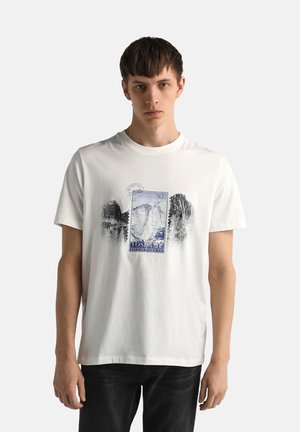 Joven vestido con una camiseta blanca con un gráfico de sellos postales temáticos de Yosemite, combinado con pantalones negros, posando frente a un fondo liso.