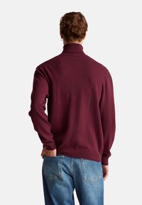 Bordowy sweter z golfem o teksturowanym wykończeniu, z długimi rękawami oraz ściągaczami na rękawach i u dołu, w zestawieniu z jasnoniebieskimi dżinsami.