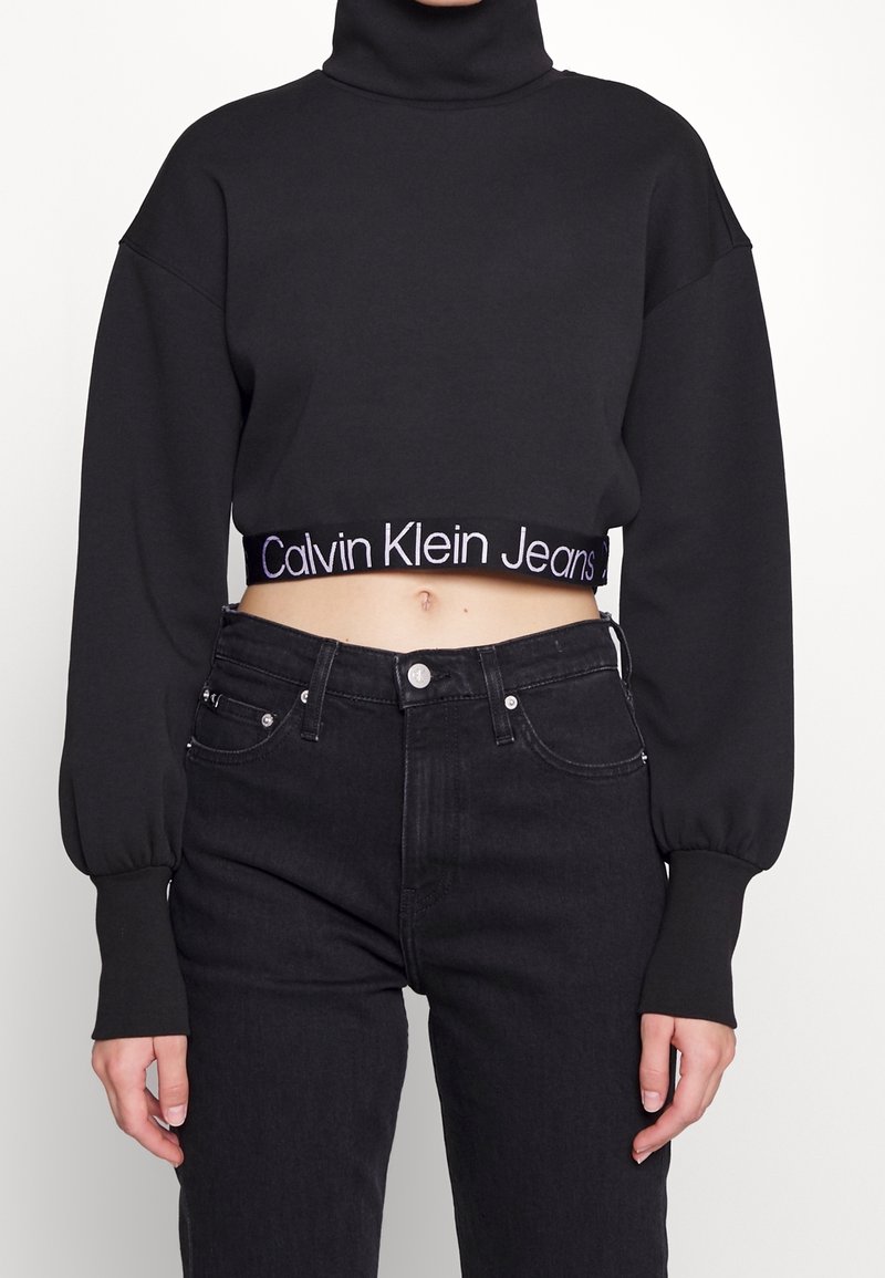 Femme portant un sweat-shirt court à col roulé noir avec "Calvin Klein Jeans" sur l'ourlet et un jean noir taille haute.