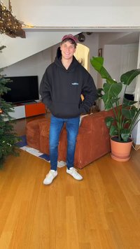 Zwarte hoodie met logo, blauwe jeans en witte sneakers. De scène bevat een plant, een bruine bank en een tv in een goed verlichte kamer.