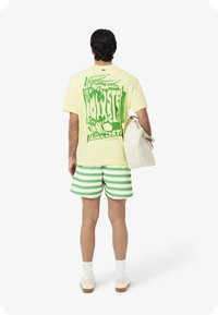 Camiseta gráfica amarilla con estampado verde, combinada con pantalones cortos a rayas verdes y blancas. Sosteniendo un bolso de color claro, de pie sobre un fondo sencillo.