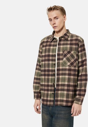 Junger Mann trägt ein grün-braun kariertes Hemd über einem weißen T-Shirt, kombiniert mit blauen Jeans, steht vor einem schlichten hellen Hintergrund.