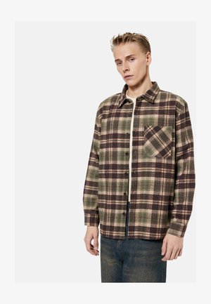 Junger Mann trägt ein grün-braun kariertes Hemd über einem weißen T-Shirt, kombiniert mit blauen Jeans, steht vor einem schlichten hellen Hintergrund.