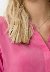 Blusa leggera e trasparente di colore rosa con scollo a V. Presenta maniche corte e una texture liscia, che si abbina a una semplice collana d'argento.