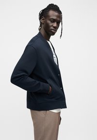 Ναυτικό μπλε ζακέτα με στρογγυλή λαιμόκοψη, κουμπιά στο μπροστινό μέρος και ριμπ μανσέτες. Φοράται πάνω από ένα λευκό γραφικό μπλουζάκι και μπεζ παντελόνια.