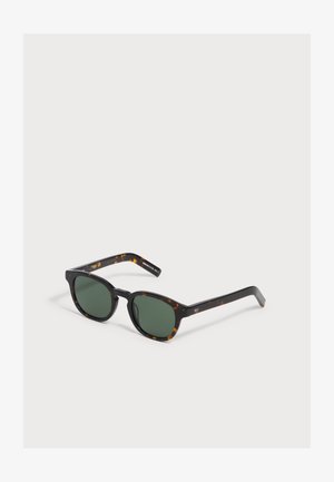 Sonnenbrille mit dunkelgrünen Gläsern und schwarzem Gestell mit braunen Schildpatt-Akzenten, auf einer weißen Oberfläche liegend.