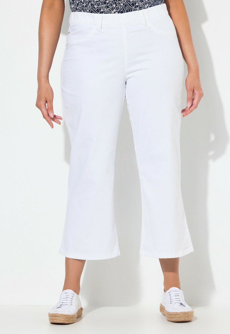 Femme portant un pantalon blanc court à jambes larges, un haut bleu marine à motifs et des baskets espadrilles blanches, debout devant un fond clair uni.