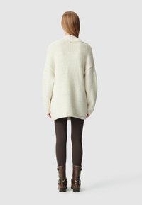 Maglione in crema extra large con colletto a coste, texture morbida e orlo asimmetrico, abbinato a leggings marroni e stivaletti alla caviglia marroni.