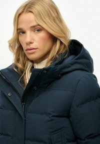 Chaqueta acolchada azul marino con capucha, que cuenta con un cierre de cremallera y botón a presión. Tiene una textura acolchada y un detalle de bolsillo lateral.