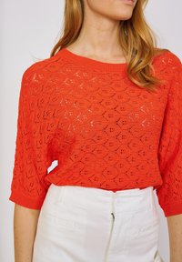 Nainen, jolla on kirkkaan oranssi neulepaita, jossa on lehtikuvio, ja valkoiset korkeavyötäröiset housut, kuvattu olkapäistä ja vyötäröstä.