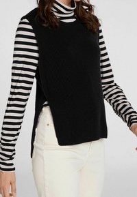 Zwarte mouwloze gebreide top over een zwart-wit gestreepte turtleneck, gecombineerd met witte broek. Zijslits in de top voor extra detail.