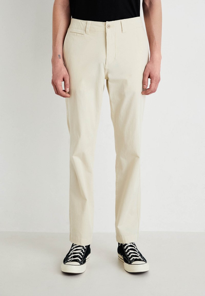Pantaloni din bumbac bej cu un design drept, având buzunare laterale și închidere cu nasture, asortați cu adidași negri cu guler înalt.