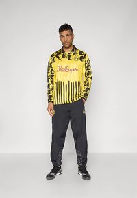 Gelbes Langarmtrikot mit markanten schwarzen Mustern und Tropfdesign, versehen mit dem "KidSuper"-Logo. Kombiniert mit schwarzen Hosen, mit strukturierten Akzenten an den Beinen.