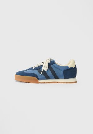 Blue and navy low-top sneaker with white laces, gum sole, and beige heel tab, shown from the side on a plain background.