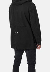 Giacca nera con cappuccio dal taglio relaxed, realizzata in tessuto liscio, con due bottoni a pressione argentati sul retro e tasche laterali nelle cuciture.