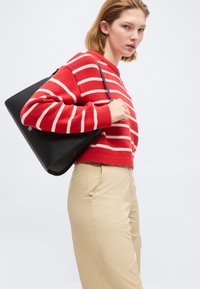 Γυναίκα που φορεί κόκκινη και λευκή ριγέ μπλούζα και μπεζ παντελόνι, κρατώντας μια μαύρη τσάντα ώμου μπροστά σε ομοιόμορφο ανοιχτό φόντο.