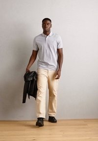 Grå polo shirt, beige bukser, sorte sko, holder en sort læderjakke. Modellen står foran en neutral baggrund med trægulv.