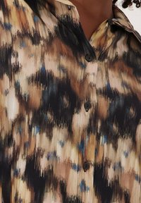 Knoopshirt in een mix van bruin, zwart en beige tinten met een gestructureerd, abstract patroon; heeft kleine zwarte knopen en een kraag.