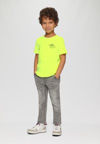 Neon gele T-shirt met korte mouwen en een dinosaurusafbeelding, gecombineerd met grijze spijkerbroeken en witte sneakers met rode accenten. Kind staat met de handen in de zakken.