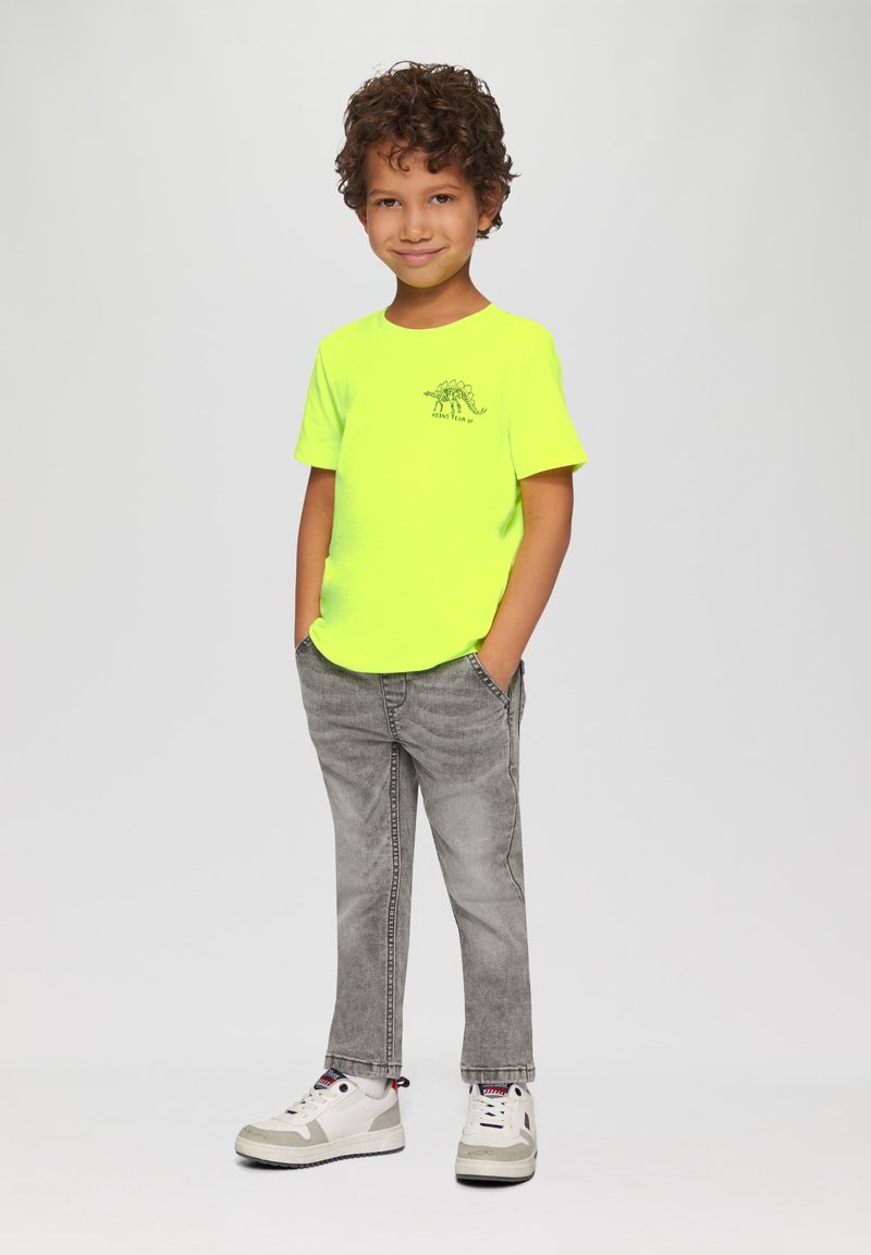 Neon gele T-shirt met korte mouwen en een dinosaurusafbeelding, gecombineerd met grijze spijkerbroeken en witte sneakers met rode accenten. Kind staat met de handen in de zakken.